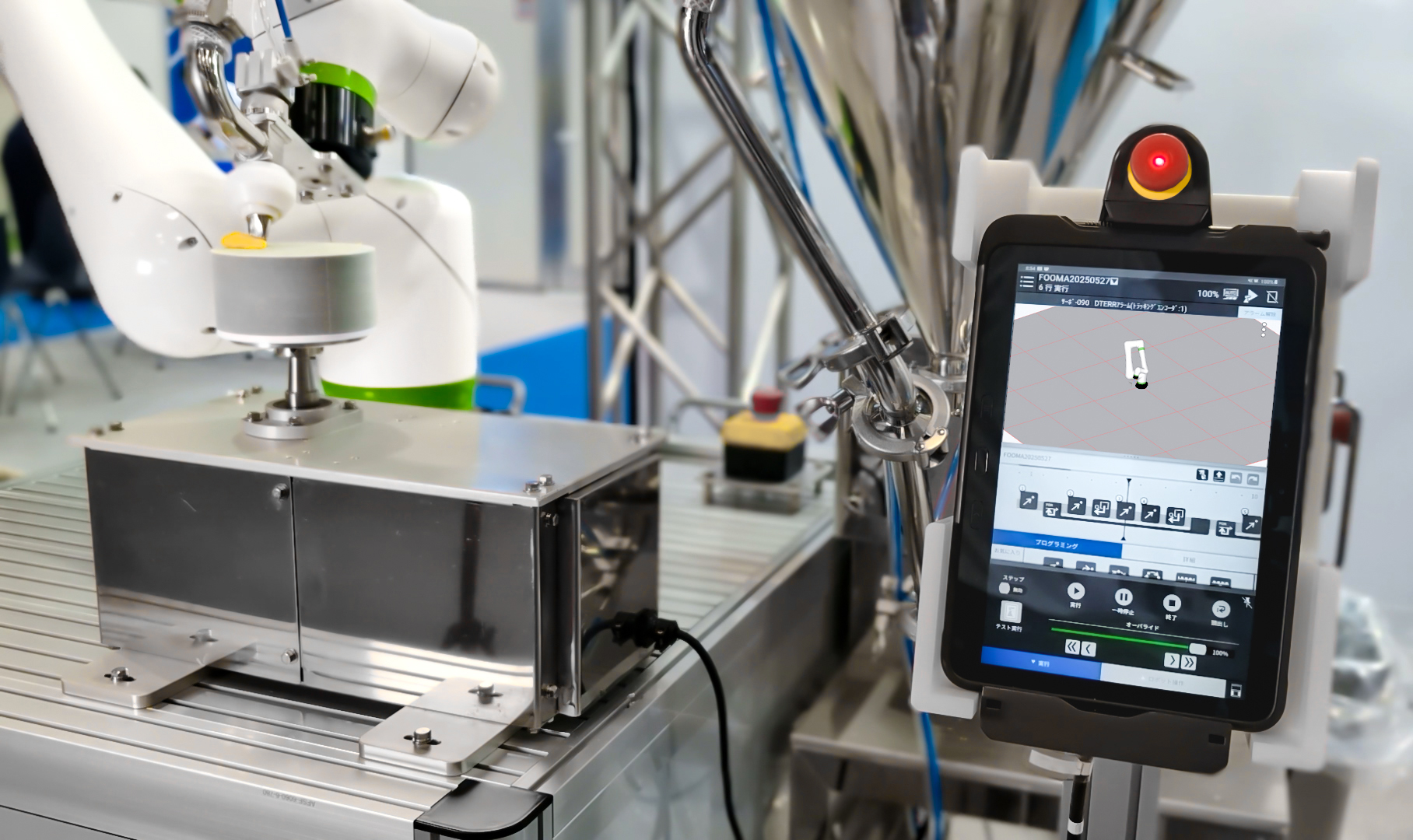 A white FANUC collaborative robot arm working at a metal bench beside a handheld teach pendant with a touchscreen interface and red emergency stop button.