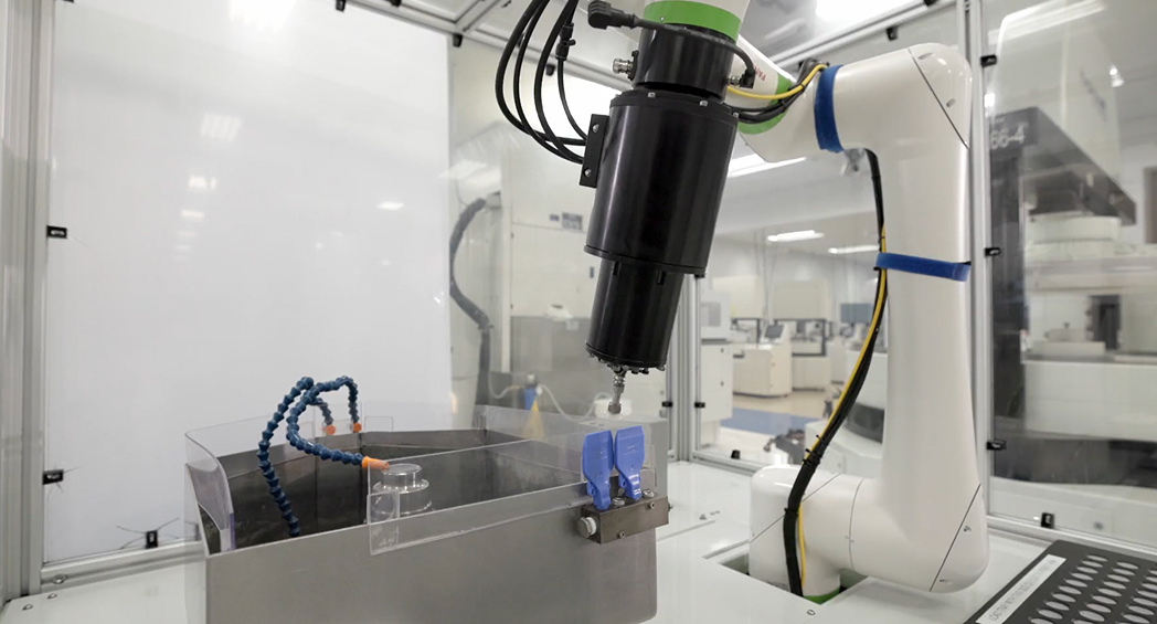 White collaborative robot arm with a black tool attachment positioned over a small metal fixture inside a factory work cell with clear plastic safety panels, other machines visible in the background.