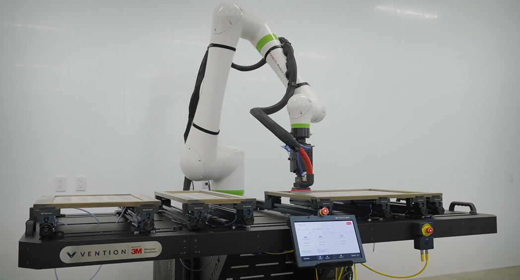 A white FANUC collaborative robot arm with a sanding attachment working on a flat panel mounted on a Vention automation platform with a touchscreen control panel.