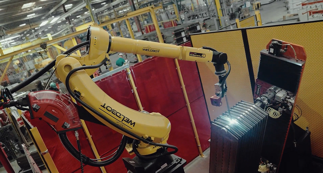 A yellow 'WELDBOT' robotic welding arm performing arc welding on stacked metal fins within a manufacturing facility, with red curtain barriers and factory equipment visible in the background.