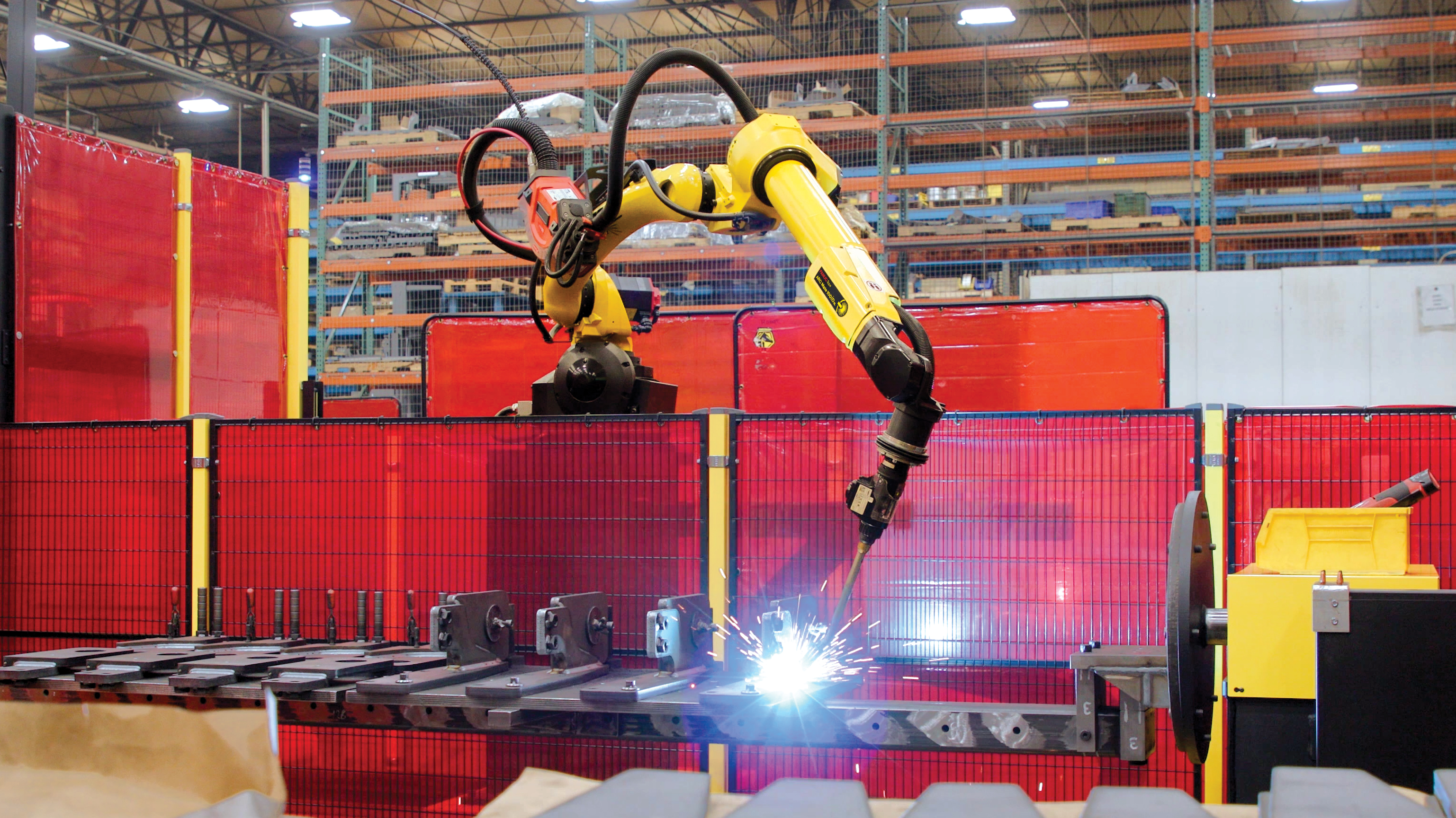 A yellow and black FANUC ARC Mate 100i welding robot arm is performing arc welding on metal parts within a red safety-fenced work cell in an industrial warehouse.