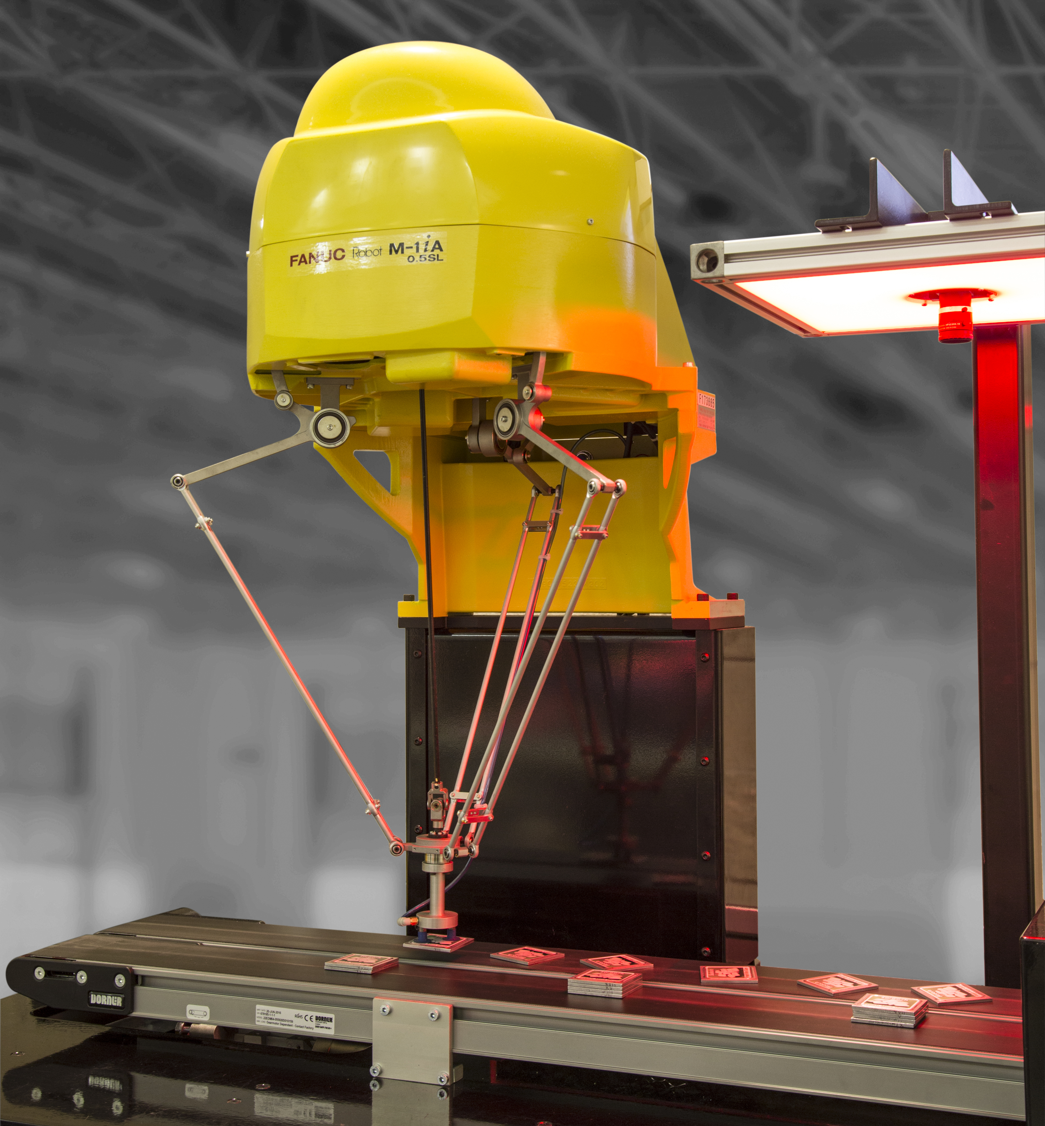 A yellow FANUC M-1iA parallel-link robot mounted above a conveyor belt, using its delta-arm mechanism to pick and place small red items under red inspection lighting.