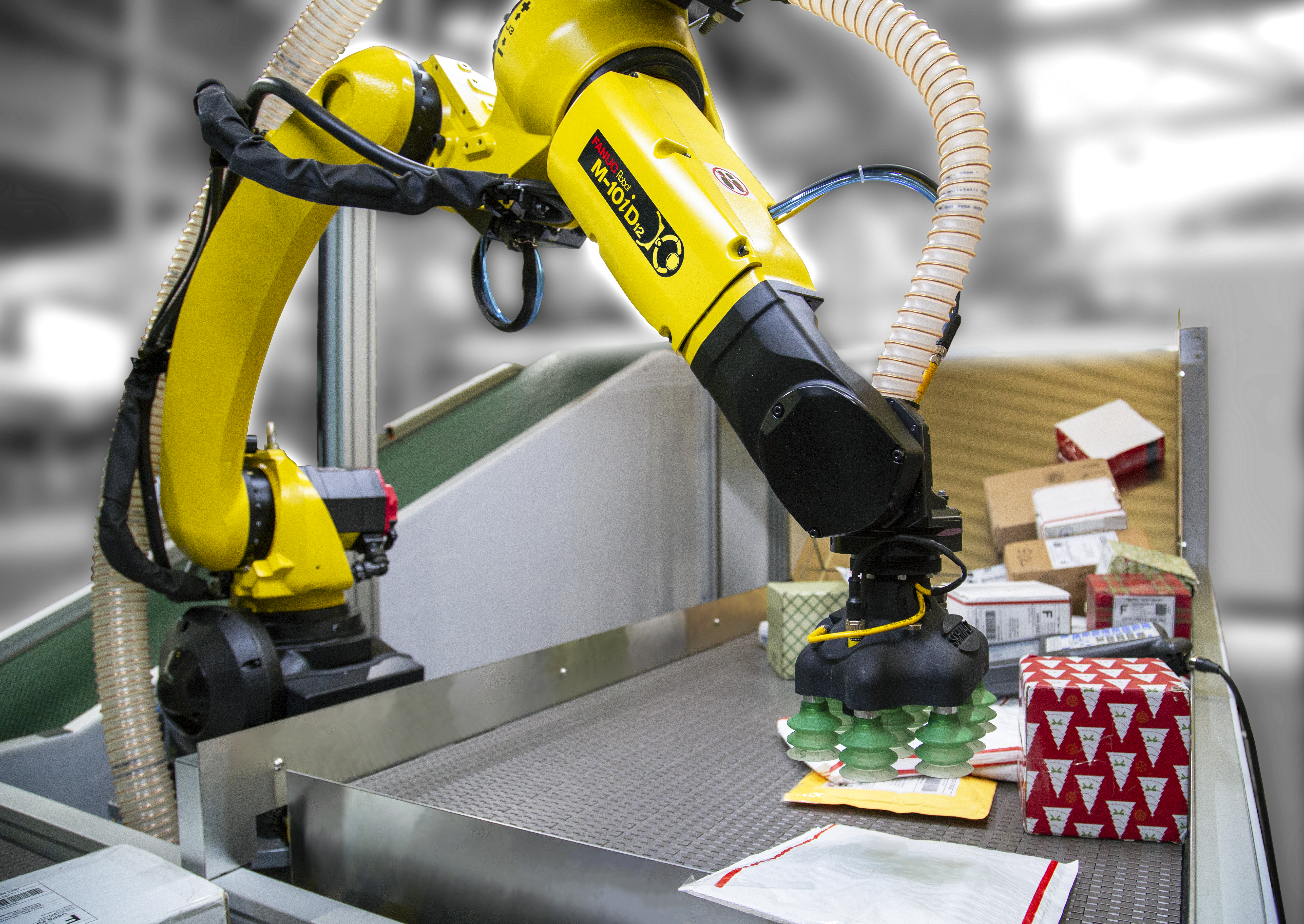 Yellow and black industrial robot arm with a suction-cup gripper picking up a package from a conveyor belt surrounded by boxes and mail parcels in a warehouse setting.