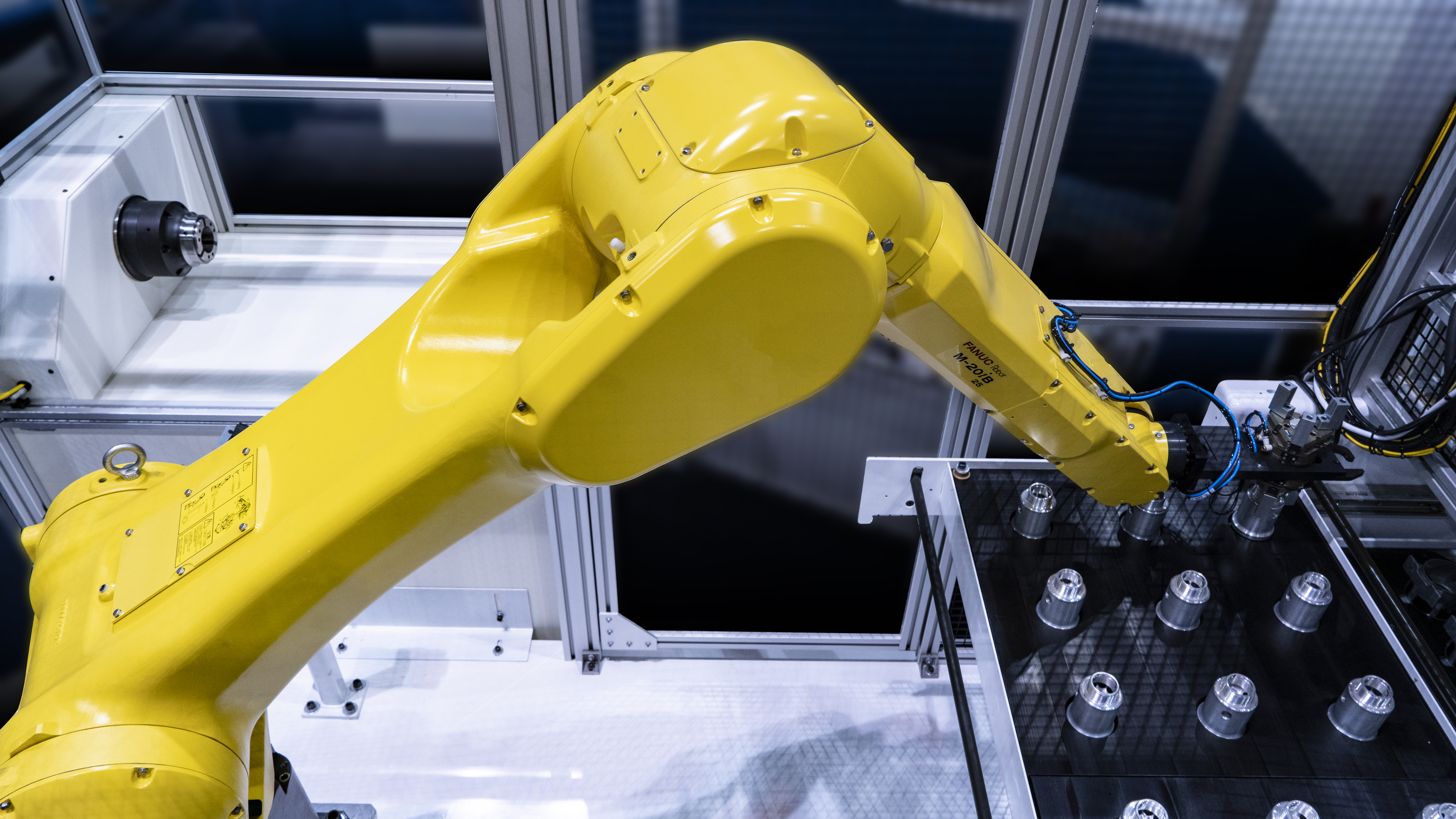 Yellow M-20iB/25 industrial robot arm inside a machining cell, reaching toward a tray of cylindrical tool holders, with an aluminum-framed enclosure in the background.