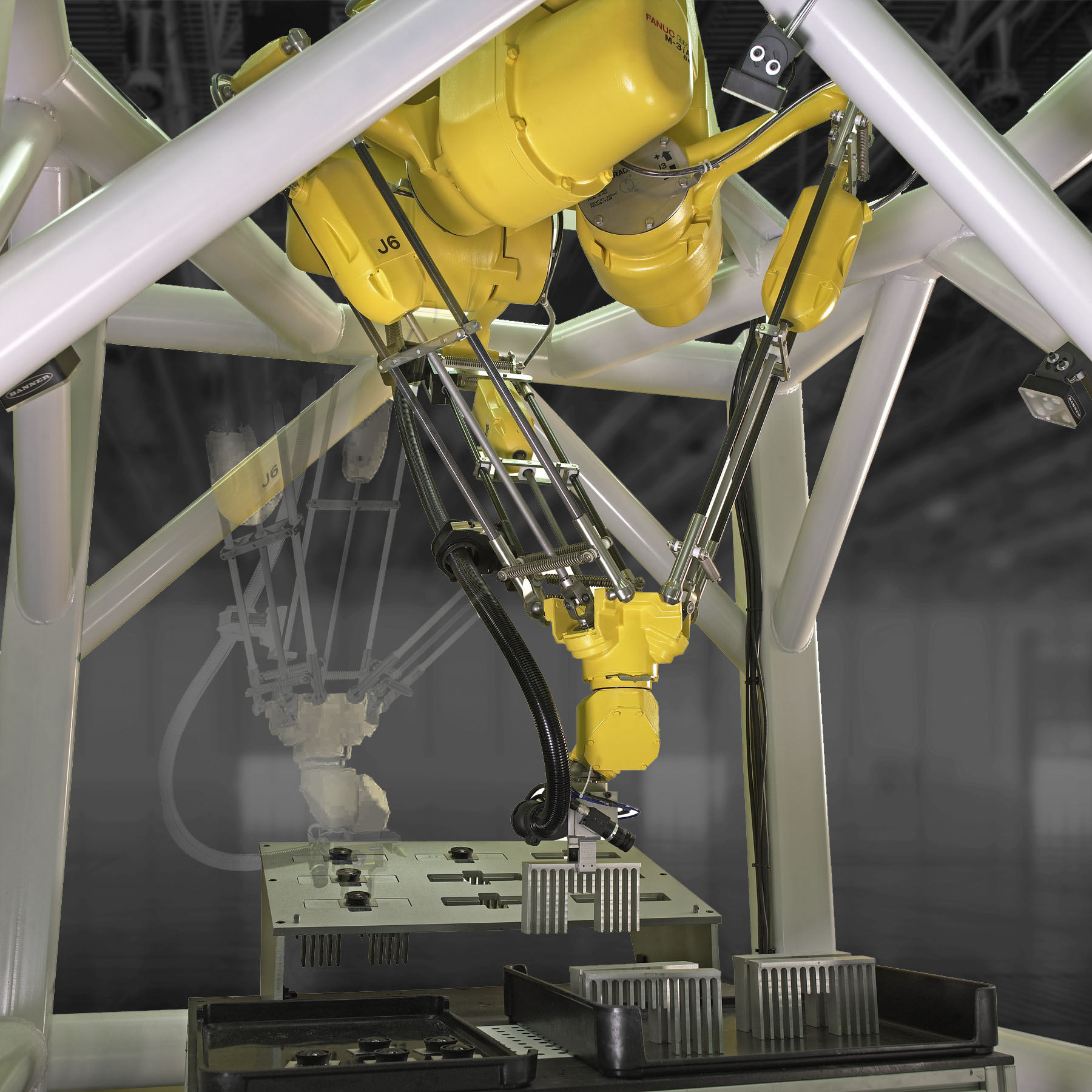 A yellow FANUC M3i A parallel-link robot mounted in a metal frame, using its articulated arm to place a component into a tray on a workstation below.