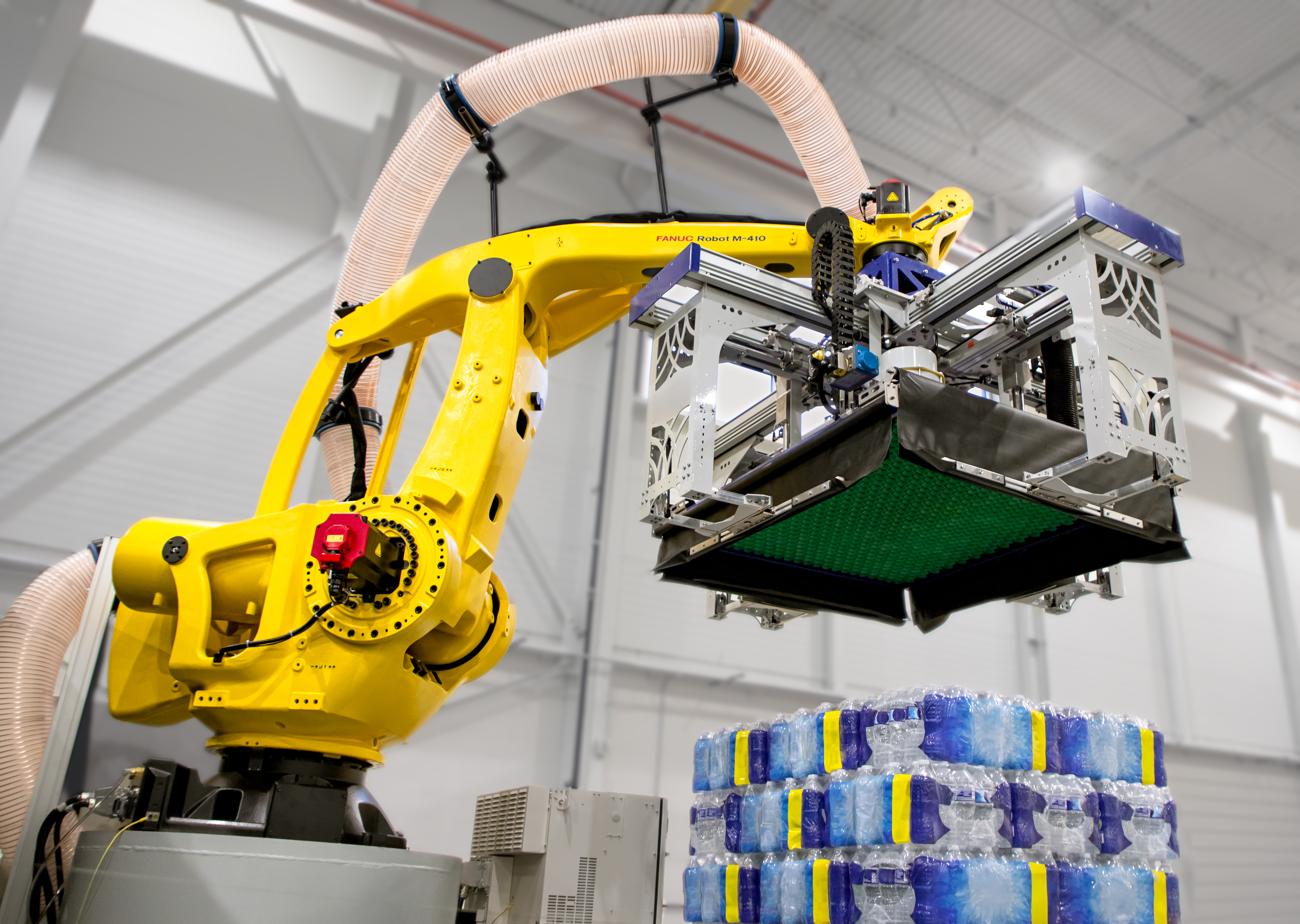 A yellow FANUC M-410iC robotic arm with a large gripping frame lifting a layer of packaged water bottles from a stacked pallet in a warehouse.