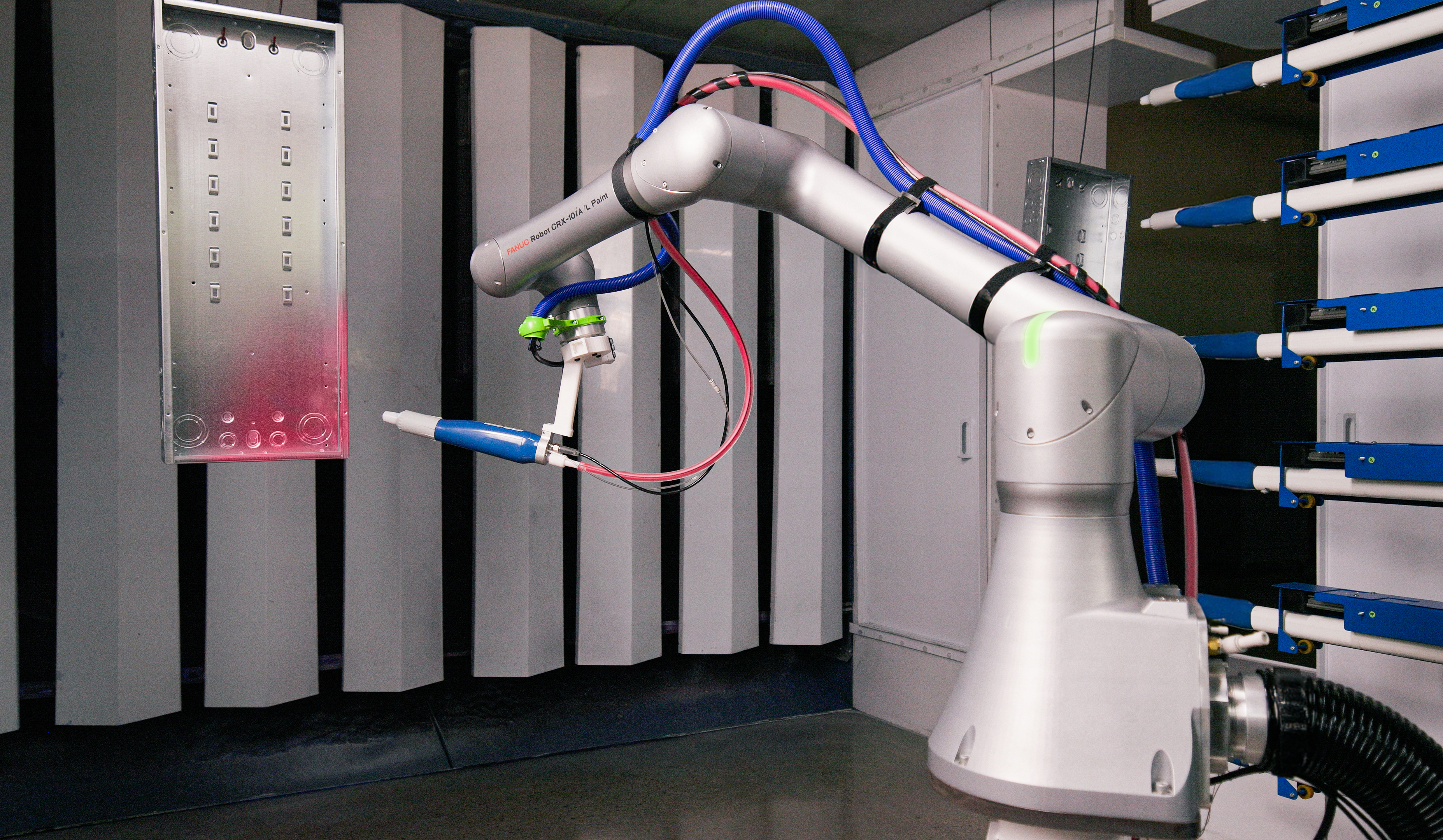 A white robotic paint-spraying arm with blue and red hoses, positioned near a slatted booth wall with a pink-tinted panel showing spray residue.