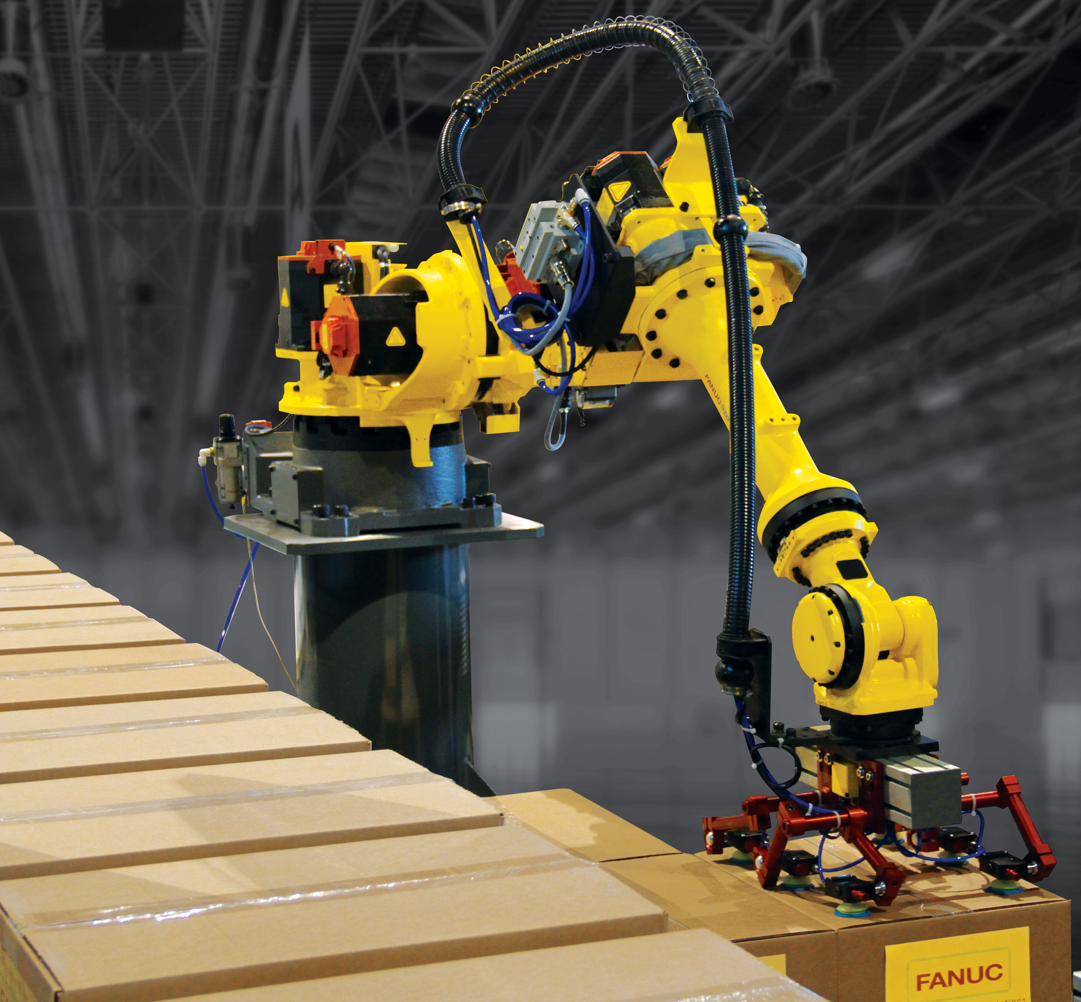 A yellow FANUC R-1000i industrial robot arm with a red vacuum suction gripper lifts a cardboard case from a conveyor line onto a stacked pallet in a warehouse.
