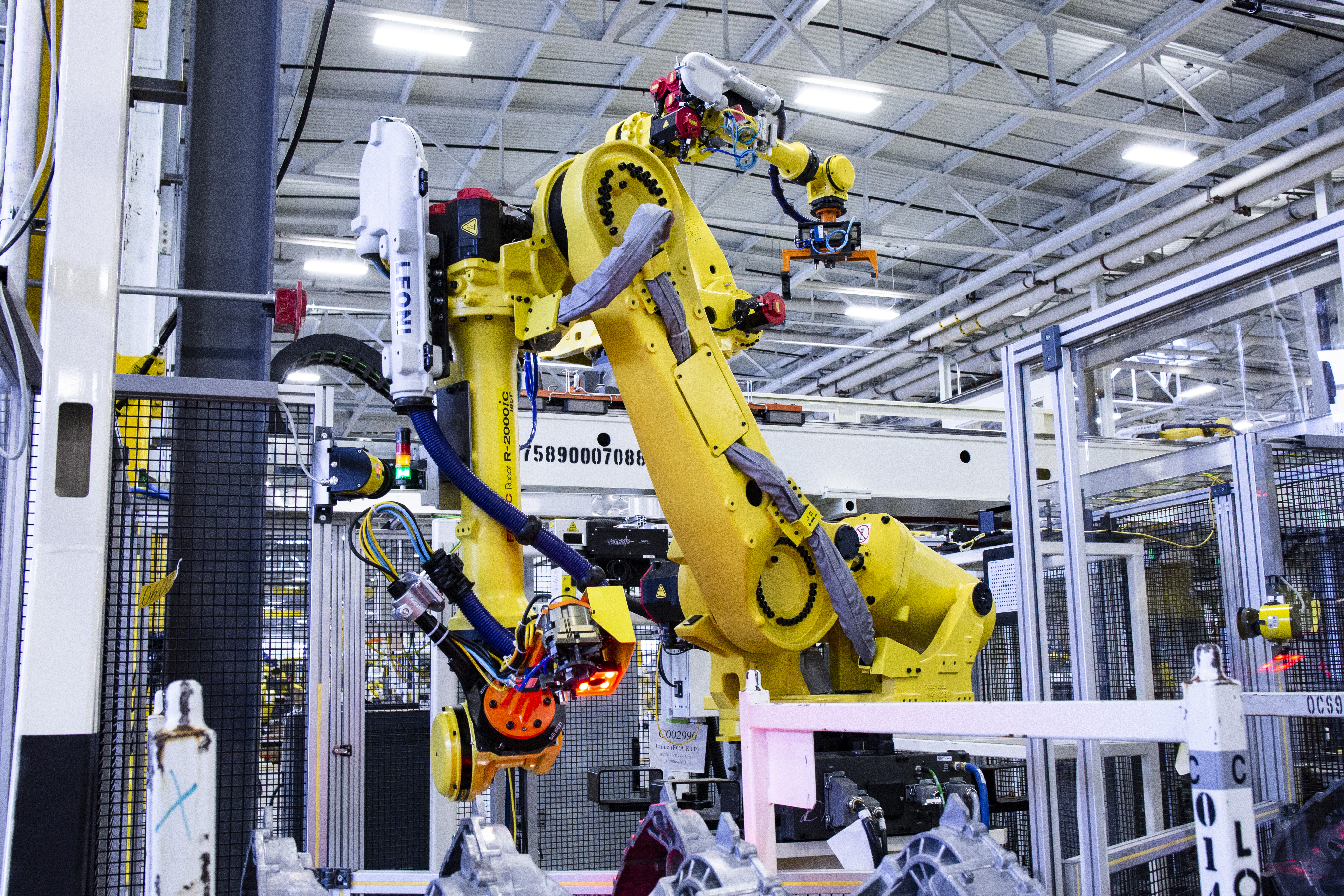 A large yellow FANUC R-2000 industrial robot arm reaching upward inside a caged automation cell on a manufacturing floor, with a smaller robot and metal parts visible nearby.