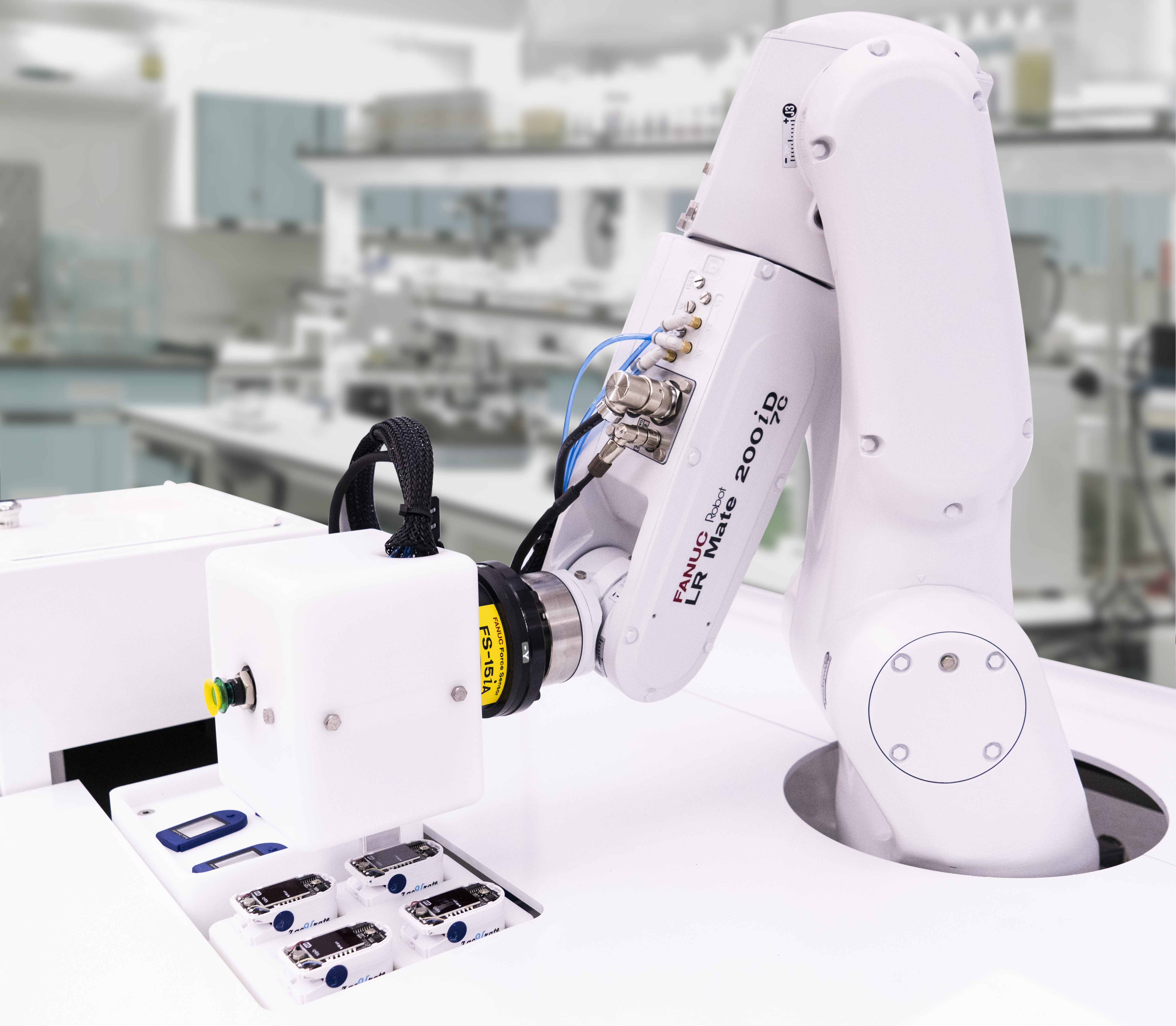 A white FANUC LR Mate 200iD robotic arm mounted on a lab bench, equipped with a vision sensor and gripper, positioned above small labeled trays in a bright laboratory setting.